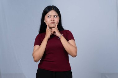 Kestane rengi tişörtlü genç bir kadın düşünceli görünüyor, parmaklarıyla yanaklarına dokunuyor.