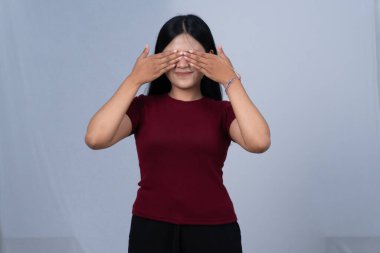 Kestane renkli tişörtlü, gülümseyen bir kadın iki eliyle gözlerini kapatıyor.