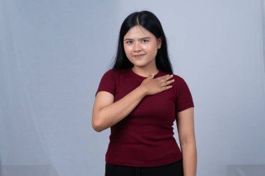 Kestane rengi tişörtlü, gülümseyen genç bir kadın elini kalbine koyar.