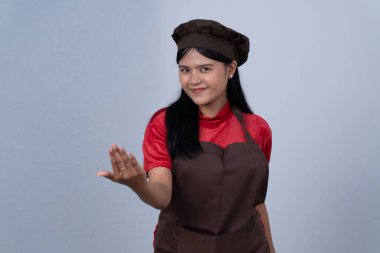 Kahverengi önlüklü ve şapkalı bir kadın şef gülümseyerek yaklaşan birine işaret eder.