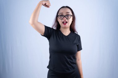 Siyah tişörtlü güçlü bir Asyalı kadın pazılarını esnetiyor ve ağzı açık bağırıyor.