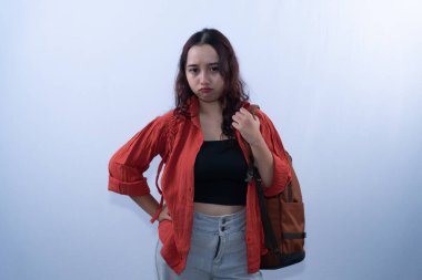 Kırmızı tişörtlü, sırt çantalı, eli kalçasında ve somurtkan Asyalı bir kadın.