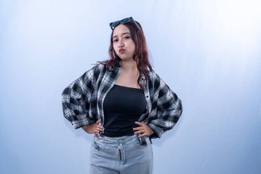 Ekose gömlekli ve güneş gözlüklü Asyalı bir kadın elleri kalçasında ve somurtkan bir ifadeyle poz veriyor.