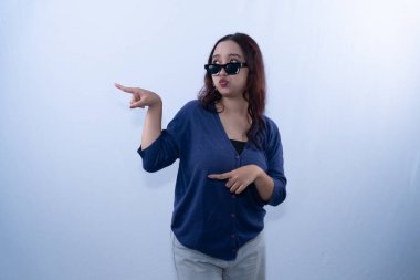 Güneş gözlüğü takan, dudaklarını büzüştüren ve beyaz bir arka plana yan bakan genç bir kadın.