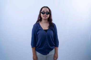 Mavi hırka, gri pantolon ve güneş gözlüğü takan genç bir kadın fotoğraf için gülümsüyor.