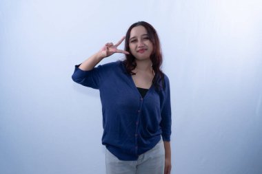 Mavi hırkalı ve gri pantolonlu gülümseyen genç bir kadın beyaz bir arka plana barış işareti yapıyor.