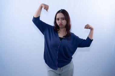 Hayal kırıklığına uğramış yüzü olan genç bir kadın, beyaz bir arka plana karşı kol kaslarını esnetirken poz verir.