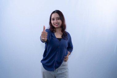 Mavi hırkalı, gülümseyen genç bir kadın beyaz bir arkaplana başparmak sallıyor.