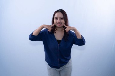 Siyah saçlı, mavi bluzlu ve gri pantolonlu gülümseyen bir kadın, mutlu poz verir.