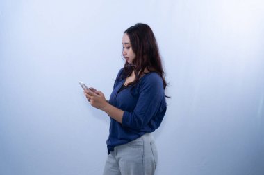 Mavi hırka giyen genç bir kadın beyaz bir arka planda akıllı telefonuna bakıyor.