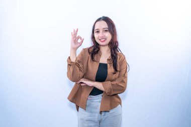 Kahverengi gömlekli, gülümseyen genç bir kadın kameraya el işareti yapıyor.