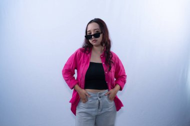 Güneş gözlüklü, pembe gömlekli ve gri pantolonlu genç bir kadın beyaz arka planda poz veriyor.