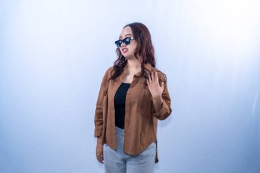 Kahverengi gömlekli, siyah güneş gözlüklü genç bir kadın eliyle poz veriyor ve hareketler yapıyor.