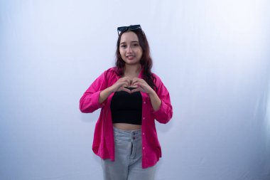 Pembe gömlekli ve gri pantolonlu gülümseyen genç bir kadın elleriyle kalp işareti yapıyor.