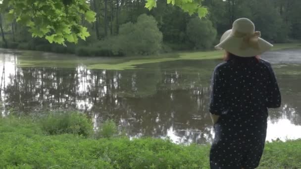 En kvinna i en hatt vid sjön i parken — Stockvideo