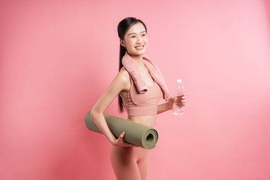 Antrenman elbiseli genç kadın pembe zemine karşı yoga minderi ve su taşıyor