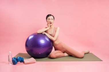 Asyalı bir kadın spor kıyafetleriyle mor bir egzersiz topuna yaslanmış şaşırmış görünüyor.