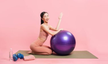 Antrenman kıyafetleri içinde genç bir kadın pembe bir arka planda gülümseyen büyük mor bir yoga topunun üzerinde dinleniyor.