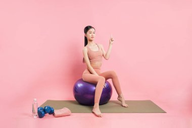 Yanında pembe bir arka planda egzersiz topunun üzerinde oturan genç bir kadın halterler, havlu ve su.