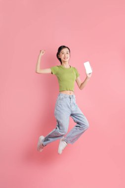 Bir stüdyo çekiminde parlak pembe bir arka plana karşı elinde telefonla sevinçle atlayan genç bir Asyalı kadın.