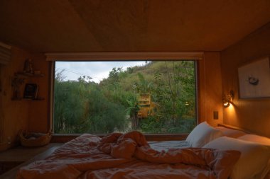 Geniş bir pencerenin önünde yatağı olan sıcak iç mekan doğal manzarayı ve yeşilliği gözler önüne seriyor.