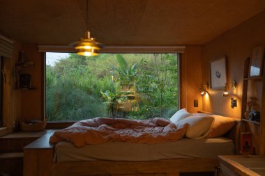 Sıcak ve davetkar bir yatak odası. Geniş bir penceresi var. Yeşil manzaralı.