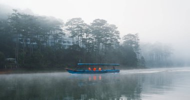 Mavi bir tekne sisli bir gölde süzülüyor. Arkasında ağaçlar beliriyor.