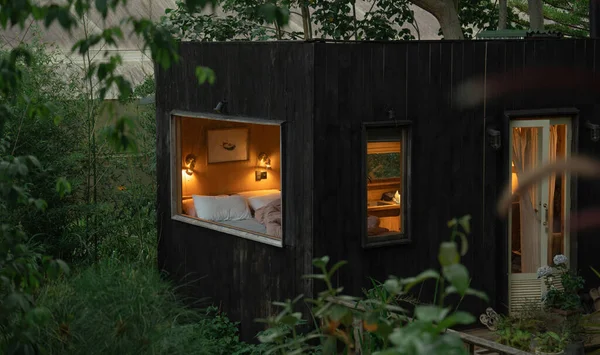 Küçük, koyu bir ahşap kulübe yemyeşil bir çevreyle harmanlanır. İçi pencerelerden sıcacık bir şekilde parlar.