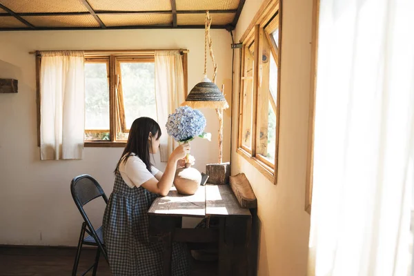 Bayan, bir vazodaki mavi bir ortanca çiçeğine hayran bir şekilde pencerenin yanında oturuyor.