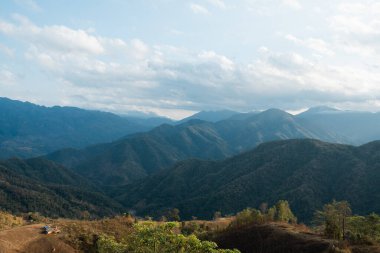 Bulutlu bir gökyüzünün altında ağaçlarla kaplı engin bir dağ silsilesi en yakın tepede küçük bir kulübe bulunur.