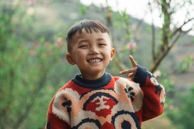 Asyalı bir çocuk, ağaçların ve pembe çiçeklerin arkasına barış işareti koyarak gülümsüyor.