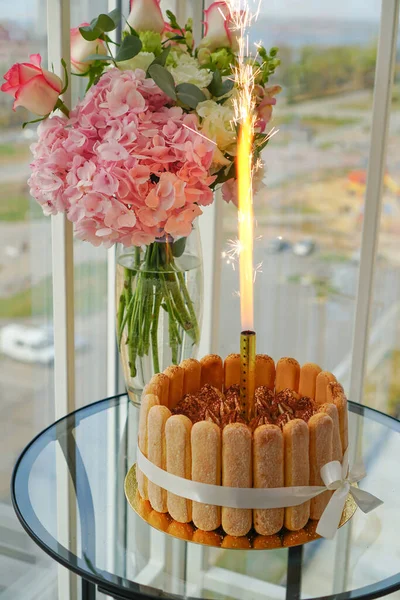 Cooking and decorating Tiramisu cake at home by a master for birthday with a lit candle. Sponge cake with flowers and cookies sprinkled with chocolate chips