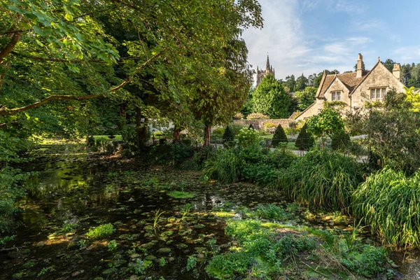 Taştan bir ev ve St. Andrew Kilisesi 'nin kulesi ile Combe Kalesi' nin yemyeşil nehir kenarı manzarası.