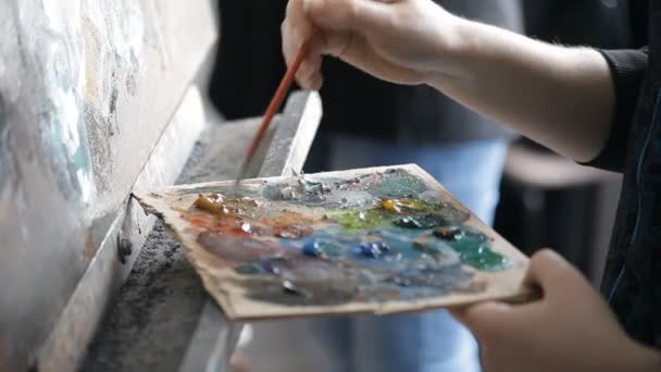 Konstnären sätter penseln till palett av färger på ett staffli — Stockvideo