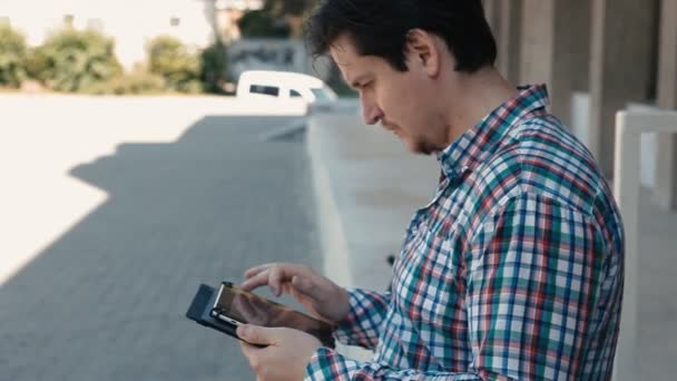 Homem em uma camisa xadrez usa o tablet ao ar livre — Vídeo de Stock