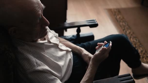 Portrait of an old man using a smartphone at home sitting in the armchair — Stock Video