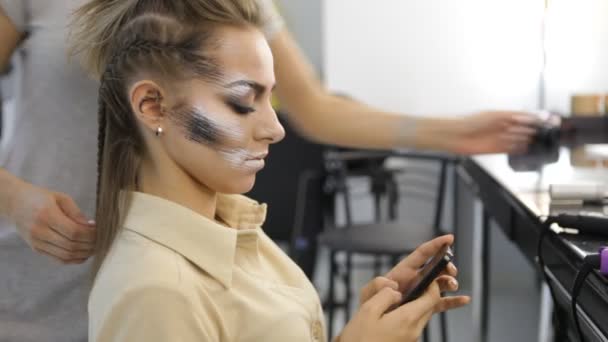 Mujer utiliza el teléfono inteligente en peluquerías mientras que el trabajo de peluquería en su cabello — Vídeos de Stock
