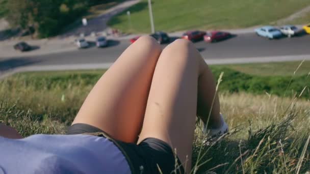 Piernas de una chica acostada en la hierba en una colina cerca de la carretera — Vídeo de stock