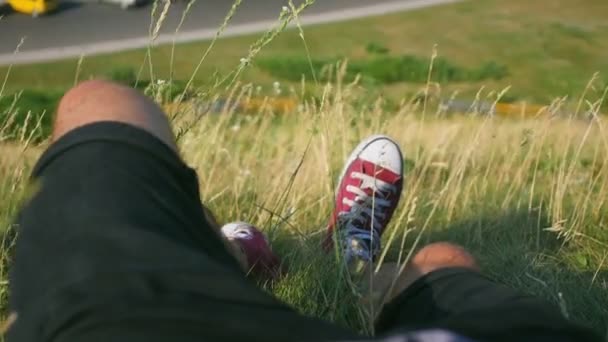 Pernas de um homem deitado na grama em uma colina perto da estrada — Vídeo de Stock