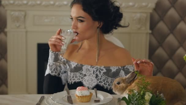 Femme en robe de soirée boit du lait et caresse un lapin assis sur la table — Video