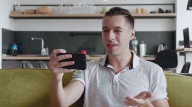 Gülümseyen genç adam evdeki kanepede otururken video görüşmesi yapıyor. Erkek portresi yaşam arka planında görüntülü konuşma yapıyor.