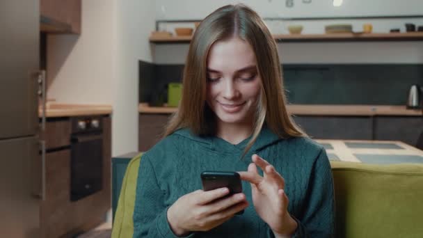 Happy young woman holding smartphone looking at screen enjoying using mobile apps having fun playing games chatting in social media sit on couch at home lifestyle — Stock Video