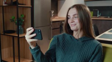 Gülümseyen, rahat genç kadın akıllı telefonu tut ve sosyal medya hikayeleri izle evdeki oturma odasındaki kanepede otur..