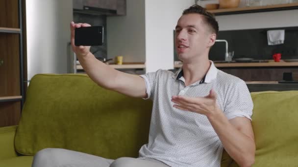 Sonriente joven haciendo videollamada mientras está sentado en un sofá en casa. Retrato de hombre haciendo un video chat en el fondo vivo — Vídeos de Stock