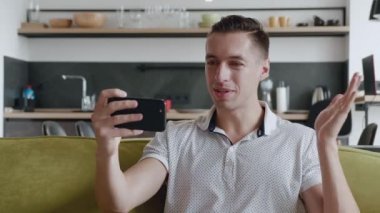 Akıllı telefonlu gülümseyen genç adam oturma odasında kanepede otururken video görüşmesi yapıyor. Mutlu adamın portresi mobil video çağrısı, evdeki koltukta oturmak, webcam görüntüsü.