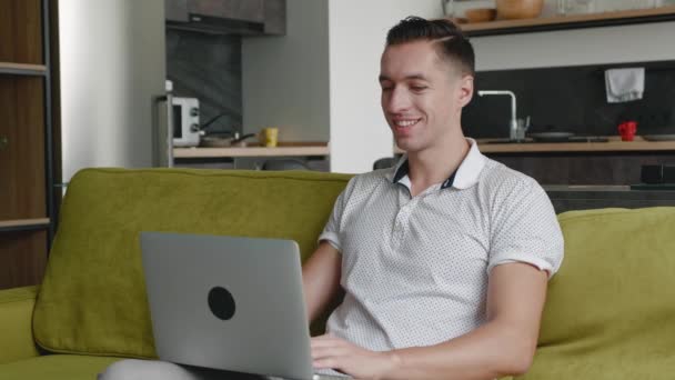 Jeune homme pigiste utilisant un ordinateur portable sur le canapé au bureau à domicile, portrait d'un homme travaillant à distance sur ordinateur dans un appartement — Video