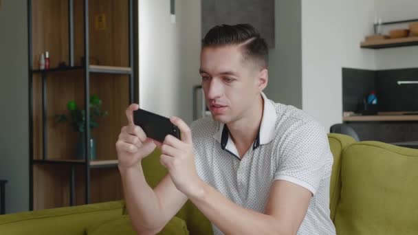 Akıllı telefonuyla oynayan genç adam heyecan verici mobil uygulaması evdeki oturma odasında kanepede otururken yumruğunu sıkarak bir yarış oyunu kazanıyor.. — Stok video