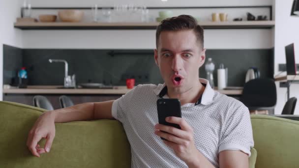 Surpreendido jovem vencedor segurar smartphone ler boas notícias espantado com celular on-line aposta lance jogo vitória em casa. Happy male look at cell phone screen overjoyed by victory success while sitting on sofa. — Vídeo de Stock