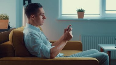 Akıllı telefonuyla oynayan genç adam heyecan verici mobil uygulaması evdeki oturma odasında kanepede otururken yumruğunu sıkarak bir yarış oyunu kazanıyor..