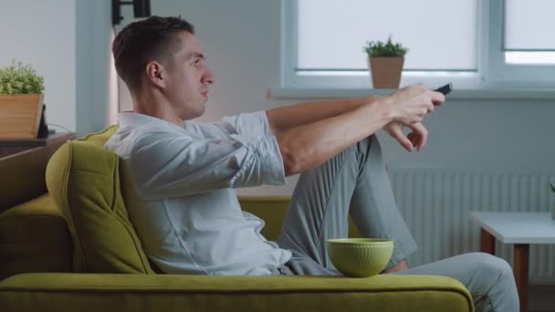 Pohledný muž se dívá na televizi a jí bramborové lupínky doma na gauči. Mladý samec sedí na pohovce a jí občerstvení, zatímco se dívá na film — Stock video